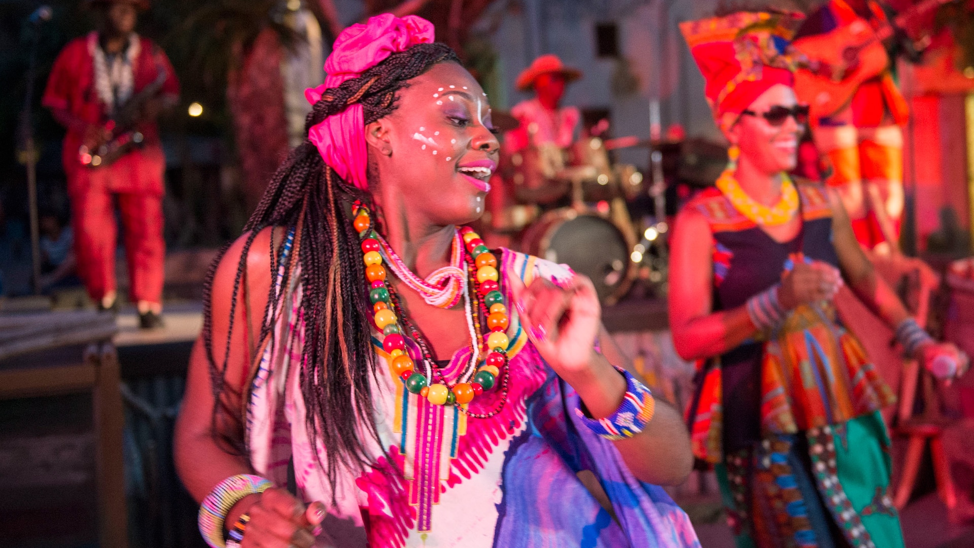 Two female performers wearing festive African style dresses and adornments dance joyously on stage as male musicians play instruments in the background