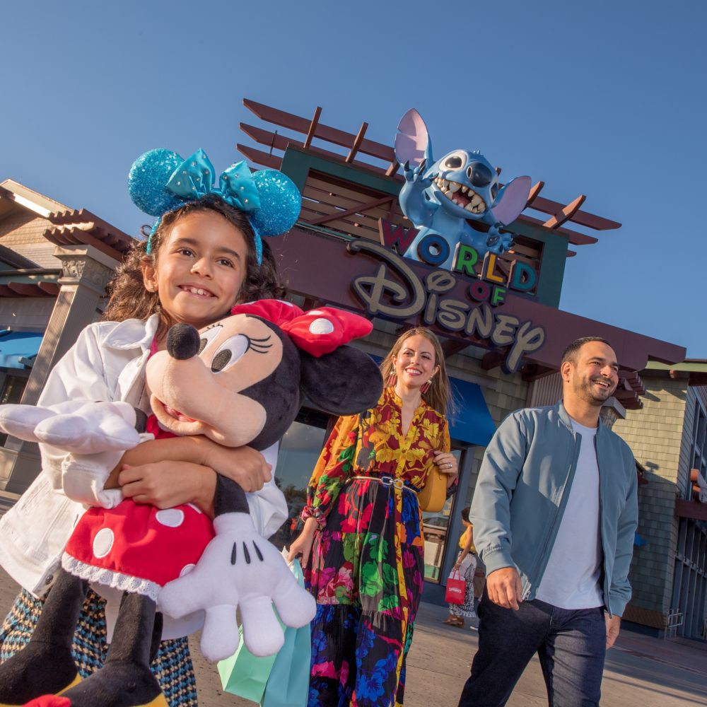 A girl hugging a large Minnie Mouse plush walking with her parents and a large Stitch figure above a sign with the words ‘World of Disney’ 