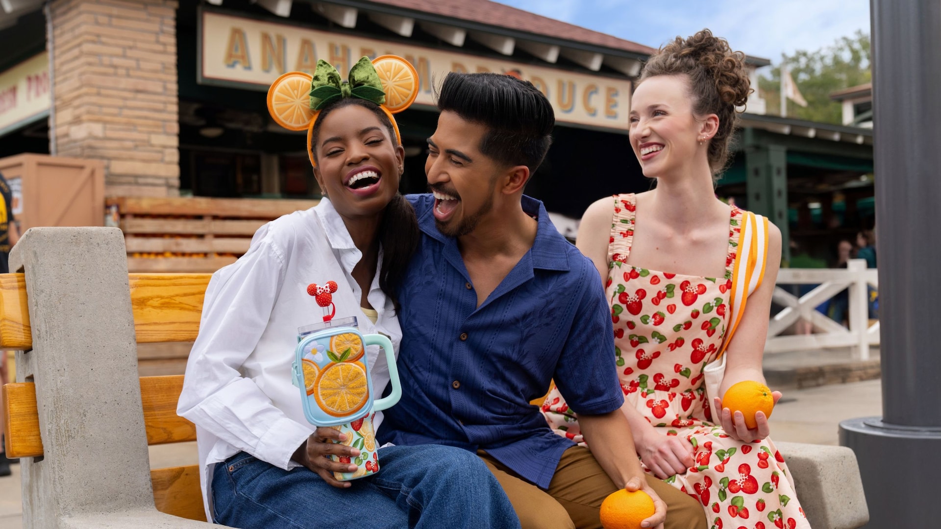 Three Guests in fruit themed apparel, including a dress, travel cup and Minnie Mouse ear headband, while sitting on a bench in front of Anaheim Produce