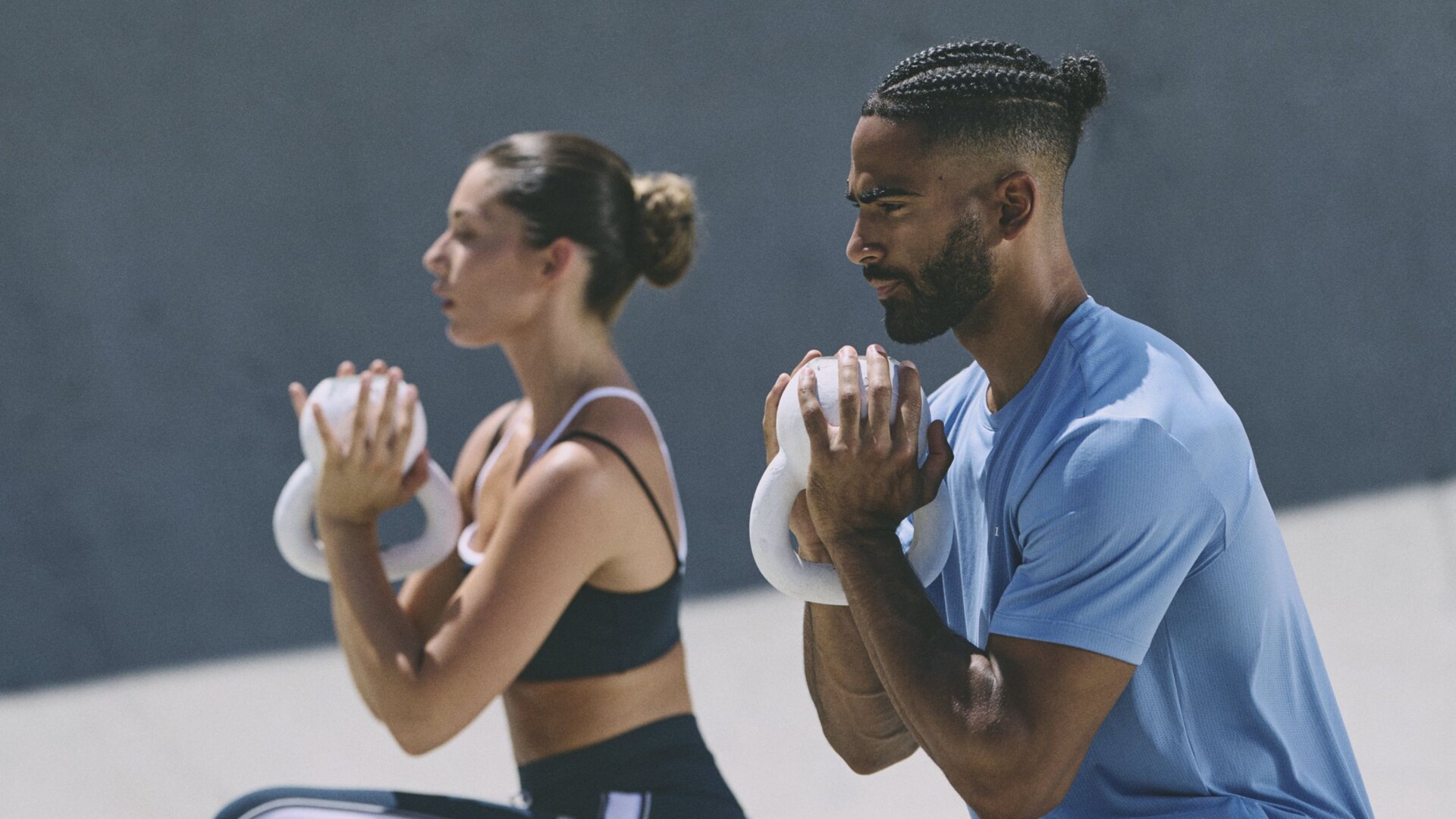 A man and woman wearing Vuori activewear while performing kneeling lunges while holding kettlebells