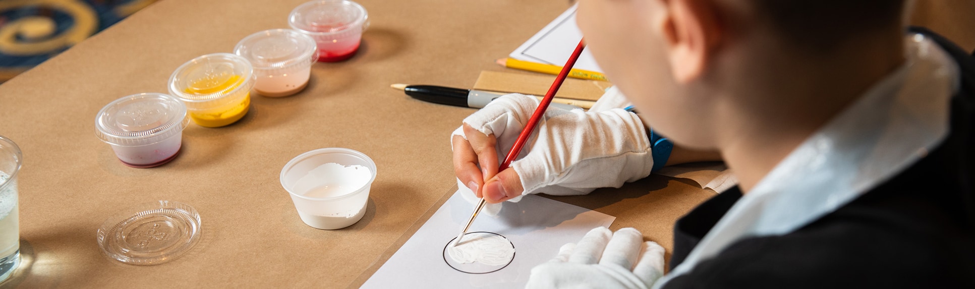 A boy wearing gloves paints inside a circle on a desk lined with art supplies
