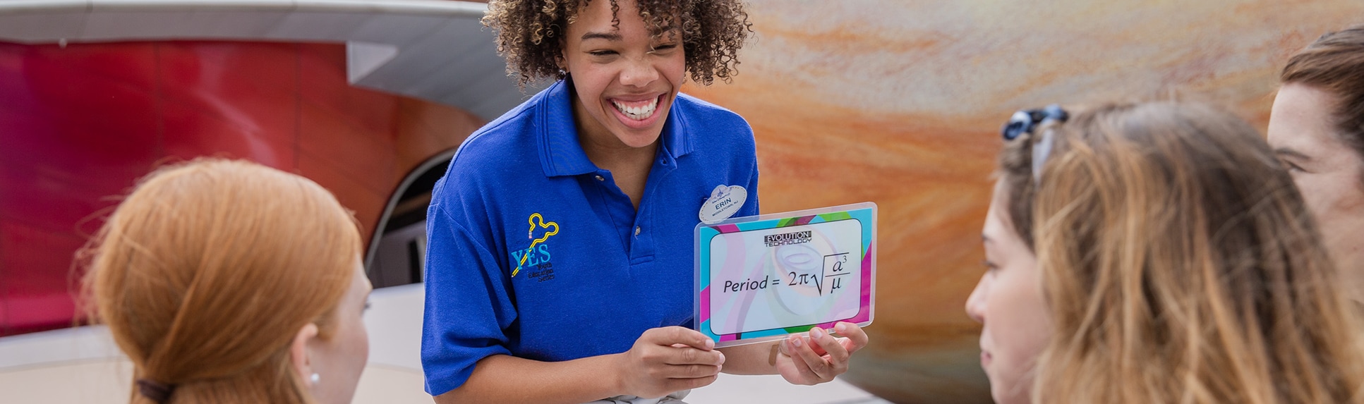 A Cast Member smiles while holding up a flashcard with a math equation to 3 teenagers