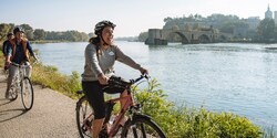 A female Adventure Guide leads a line of helmet-clad bicyclists on a path along the Rhône River past the remains of the arched Pont d’Avignon bridge A female Adventure Guide leads a line of helmet-clad bicyclists on a path along the Rhône River past the remains of the arched Pont d’Avignon bridge