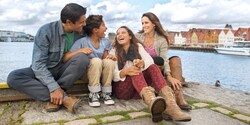 A laughing family sits together on the edge of a river, with old buildings lining the shore A laughing family sits together on the edge of a river, with old buildings lining the shore