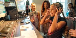 A girl with earphones sits in front of a microphone at a radio control booth, with others watching A girl with earphones sits in front of a microphone at a radio control booth, with others watching