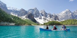 Three people in a canoe paddle across calm water with a backdrop of snow-capped mountains