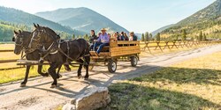 A horse and wagon, filled with people, travel down a dirt road along a wooden fence in valley between nearby mountains