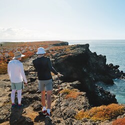 Two people wearing hats hike along a rocky clifftop high above the sea