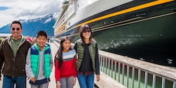 A family of 4 walk on a walkway next to a docked Disney Cruise Line cruise ship with mountains in the background A family of 4 walk on a walkway next to a docked Disney Cruise Line cruise ship with mountains in the background