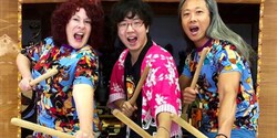 Three people with mouths agape hold drum sticks and pose by a large taiko drum