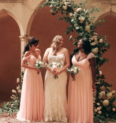 A bride standing in between 2 bridesmaids while holding bouquets in front of a floral arch