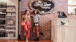 Dos mujeres jóvenes con bolsas temáticas de Disney están paradas contra una pared de ladrillos cerca de los escaparates y el mostrador de la caja registradora de la tienda