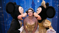 A smiling young woman in a quinceñera gown and tiara bends to let Mickey and Minnie Mouse plant a kiss on each of her cheeks