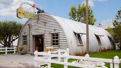 Un Cobertizo prefabricado de metal corrugado, y algo oxidado, con cerca de madera blanca, buzón, asta y un letrero que se parece a un arma de artillería pesada disparando, que dice “Sarges Surplus Hut”