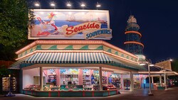 An open-air shop selling merchandise that includes sunglasses, Mickey Mouse ear hats, novelty gloves, pins, toys and snacks, beneath a lighthouse-like tower and a huge billboard sign with an illustration of a crab, a bucket of seashells, a beach, an ocean pier and the words "Seaside Souvenirs"