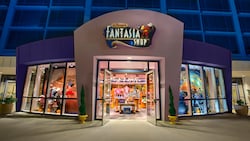 A shop at the base of a modern hotel tower with a sign featuring Mickey Mouse that reads 'Disney's Fantasia Shop' and merchandise that includes Disney-themed apparel, dolls and toys