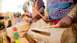 A row of percussionists drum, led by one drumming a hallow log with drum sticks