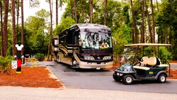 Golf cart parked in front of a gleaming motor coach parked at a wooded RV campsite