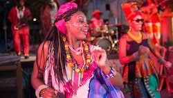 Two female performers wearing festive African style dresses and adornments dance joyously on stage as male musicians play instruments in the background