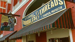 A sign above a red and green-striped awning on a red clapboard building that reads 'On the Boardwalk, Thimbles & Threads, F. Thompson, Proprietor'