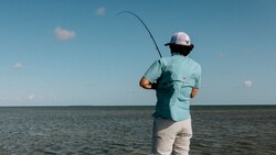 Person in outdoor apparel fishing in shallow coastal waters under a clear sky