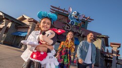 A girl hugging a large Minnie Mouse plush walking with her parents and a large Stitch figure above a sign with the words ‘World of Disney’ 