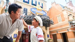 A young Guest holding a Remy plushie up to his father’s shoulder in front of Remy’s Ratatouille Adventure at Epcot
