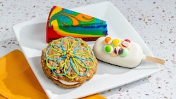 A plate featuring a slice of rainbow cheesecake next to a cake pop and a oatmeal cookie sandwich
