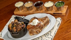 A 14 ounce dry aged grilled Duroc pork chop and roasted Brussels sprouts on a plate with a flight of 5 sauces from Le Cellier Steakhouse