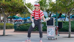 A man in a striped shirt, plaid pants and wide suspenders juggles 3 clubs and teeters on a balance board