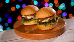 Two beef sliders topped with melted cheese, caramelized onions and leafy greens on a wooden serving board 