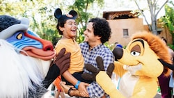 Um menino com orelhas do Mickey Mouse sendo carregado por seu pai enquanto brinca com Rafiki e Shenzi do filme The Lion King da Disney