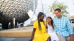 A mom, dad and their daughter laugh while sitting at the fountain in front of Spaceship Earth