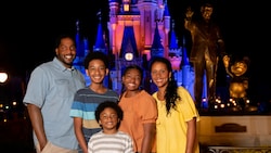 A mom, dad and 3 kids pose for a picture at night in front of the Partners Statue and Cinderella Castle