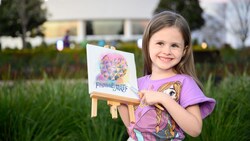A child holding a small easel with art that says ‘Epcot International Festival of the Arts’
