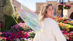 A Guest posing with a fairy wings Disney Photopass Magic Shot overlay while standing in front of a garden with flowers and a topiary