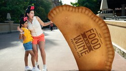 Two Guests standing next to a sign resembling a giant empanada with the Epcot International Food & Wine Festival logo
