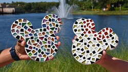 Dos personas sostienen piezas de arte en mosaico en forma de las cabezas de Mickey y Minnie frente a un lago con una fuente