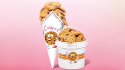 A paper cone and a bucket, each with a smiling cookie logo, overflowing with chocolate chip cookies