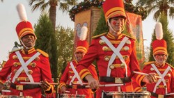 4 toy drummers in festive costumes and tall hats playing drums as they march on the street