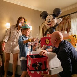 A family in a Disneyland Resort hotel room opening a Mickey Mouse birthday welcome basket featuring a Mickey Mouse ear hat and balloon 	