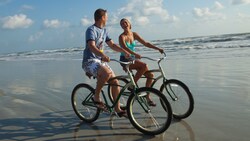 A man and a woman ride bicycles on the beach's wet sand