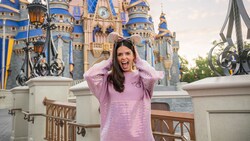 A woman wearing a shimmering sweatshirt and a sequined Minnie Mouse ear headband, standing near Cinderella Castle at Magic Kingdom park
