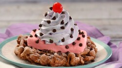 A funnel cake topped with whipped cream, chocolate chips and a cherry