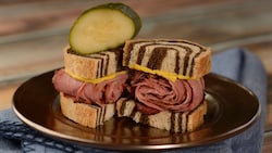 2 small pastrami sandwiches, featuring sliced pastrami between slices of rye bread, served with a pickle slice