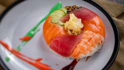 A large piece of sushi in the shape of a donut, featuring shrimp, salmon, tuna, cucumbers, sesame seeds and rice