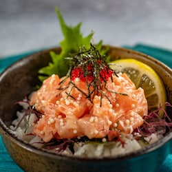 A bowl of spicy salmon served over rice, garnished with a shiso leaf and a lemon wedge