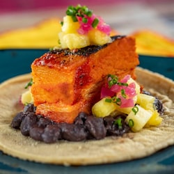 A slice of seared pork belly served with black beans and pineapple chunks, served atop a corn tortilla
