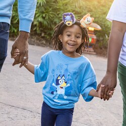 A child wearing a Bluey shirt and a Bluey and Bingo character headband walks hand in hand with 2 adults