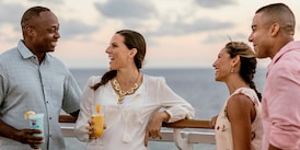 Two couples, with drinks in hand, talk while leaning on the railing of a deck aboard a ship at sea